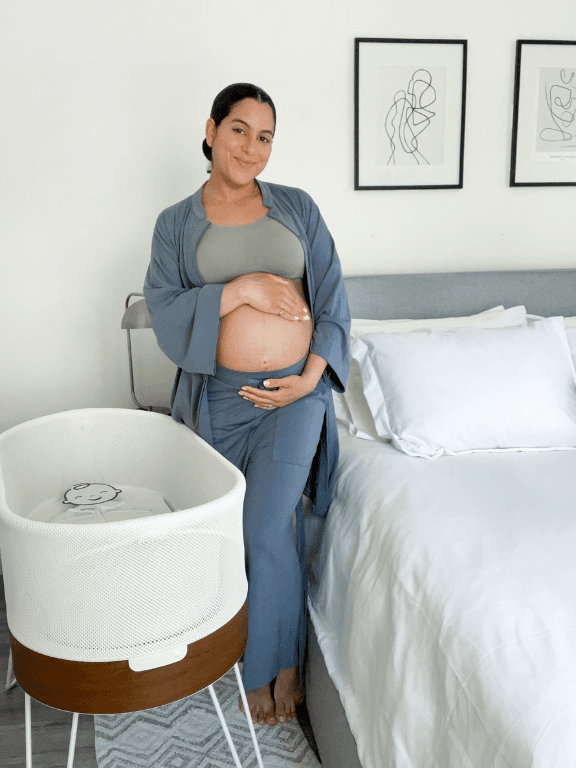 A smiling pregnant woman holds her belly next to the SNOO bassinet