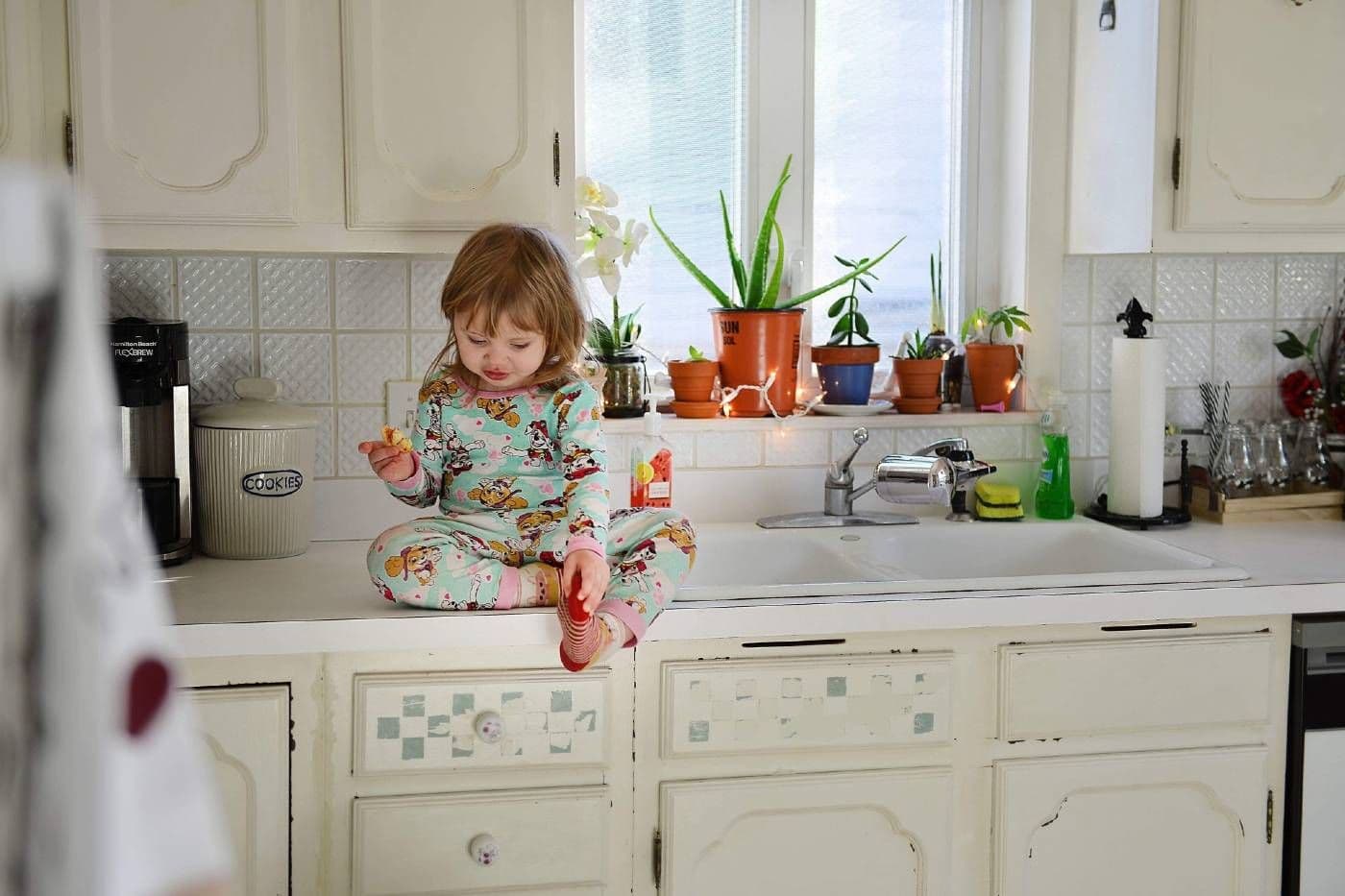A toddler girl sits on the kitchen counter and eats a snack in her pyjamas