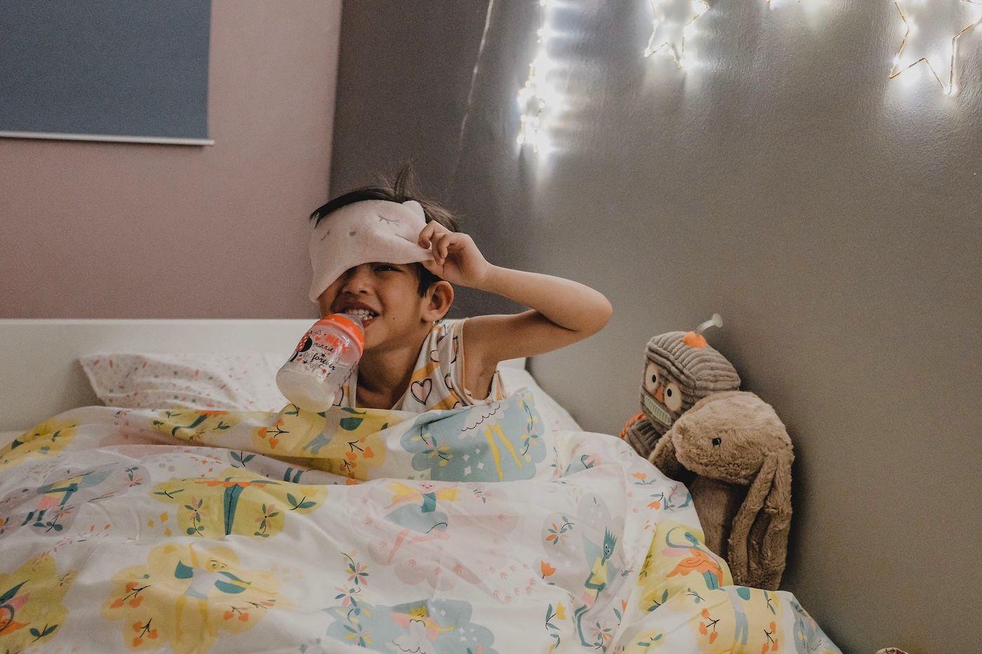 A toddler is in bed drinking a milk and wearing a sleep mask